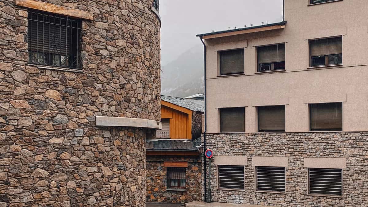 Rue d'Andorre-la-Vieille bordée de façades en pierre sèche et de toits d'ardoise, un versant boisé dans la brume au fond